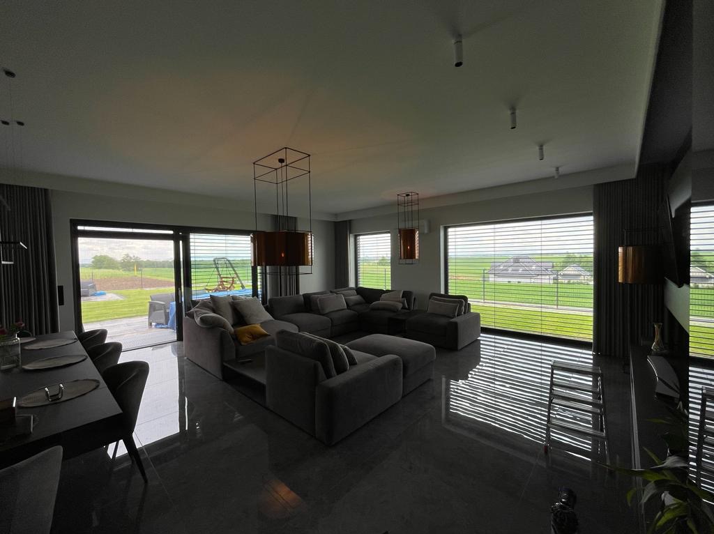 Przestronny salon z szarą, modułową sofą, wiszącymi lampami o geometrycznych kształtach i dużymi oknami z widokiem na zielony krajobraz za oknem, odbijający się w polerowanej podłodze.