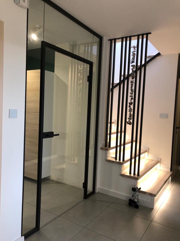 Nowoczesne wnętrze z przeszkloną ścianą z czarnymi ramami, drewnianymi schodami z czarną balustradą i dekoracją świąteczną, oraz pluszową zebrą na podłodze z szarych płytek.