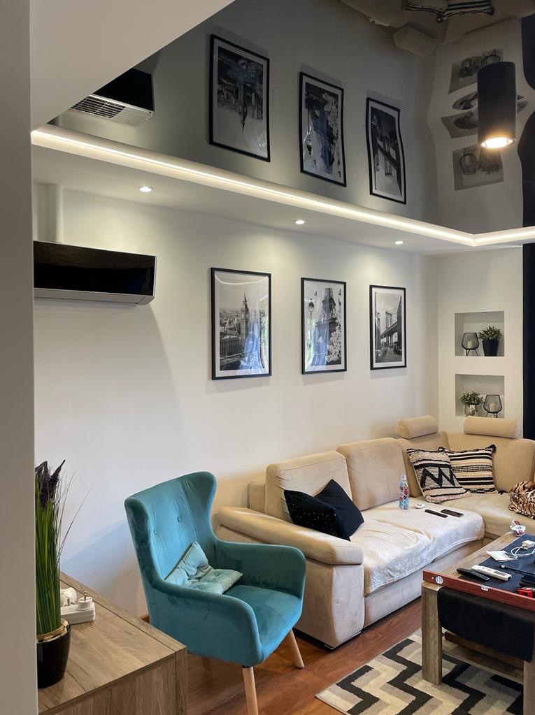 Jasne wnętrze salonu z turkusowym fotelem, beżową sofą, drewnianą podłogą i czarno-białymi obrazami na ścianie, z widocznym klimatyzatorem i oświetleniem LED.