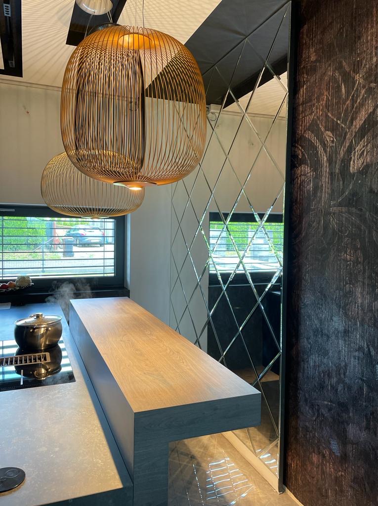 Nowoczesna kuchnia z wyspą, na której stoi garnek z parującym jedzeniem, oświetlona złotymi lampami wiszącymi, z lustrzaną ścianą w kształcie rombów i ciemną ścianą z motywem roślinnym.