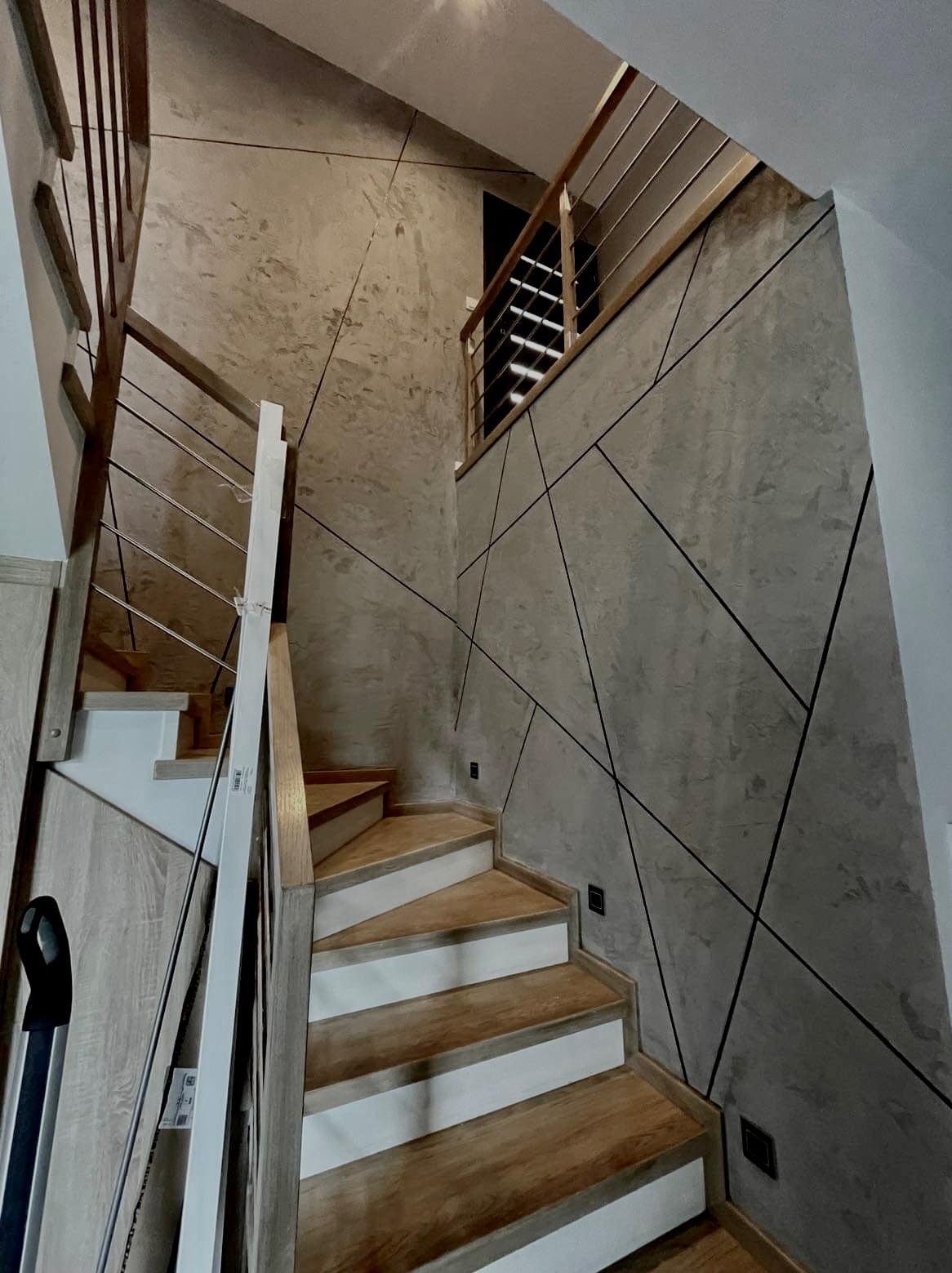 Wnętrze klatki schodowej z drewnianymi schodami z białymi podstopnicami, szare ściany z geometrycznym wzorem z czarnych linii i nowoczesną metalową balustradą.