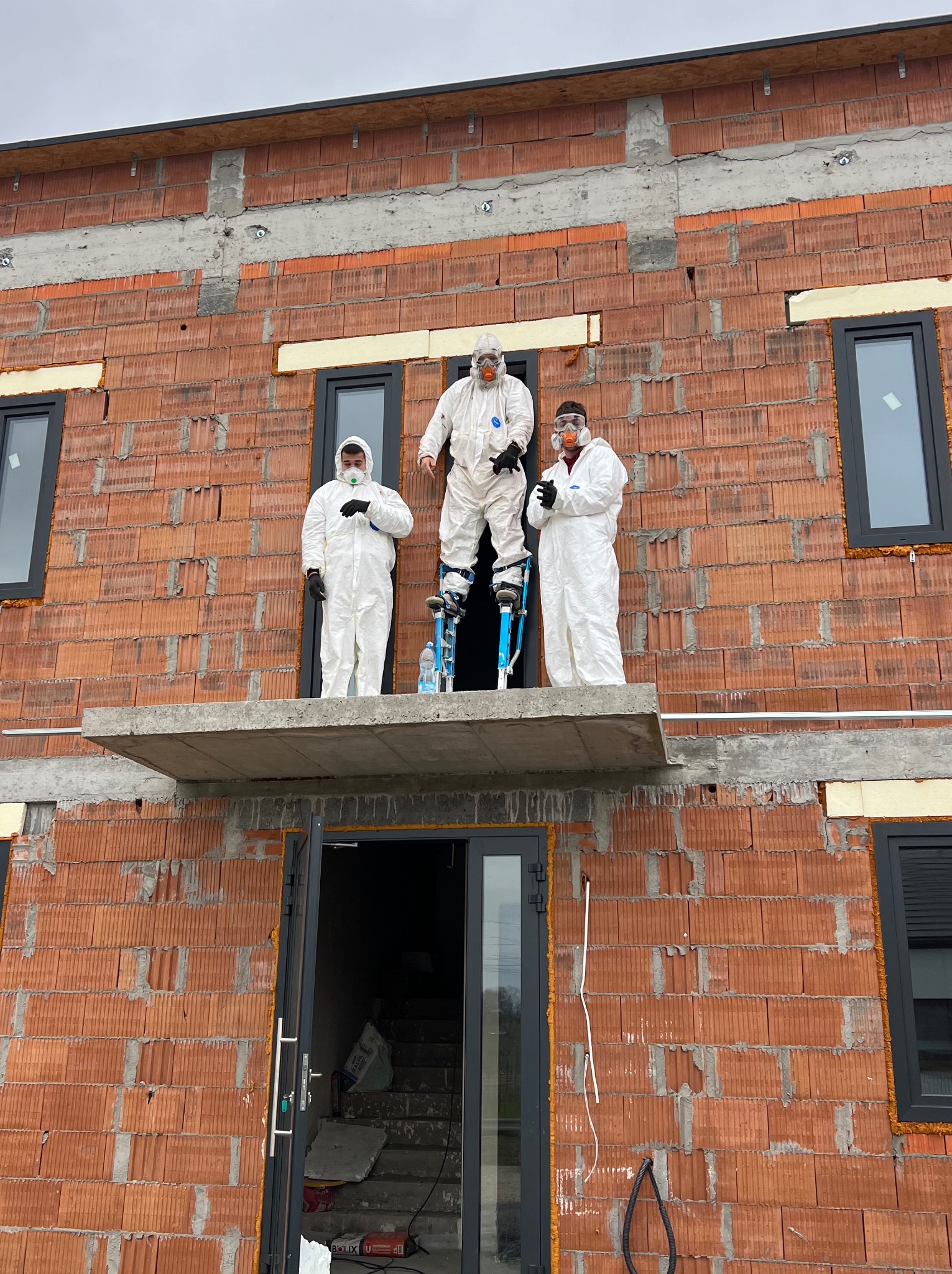 Trzech pracowników w białych kombinezonach ochronnych i maskach, jeden na szczudłach, na tle surowej ściany budynku z czerwonej cegły z widocznymi oknami i betonowymi elementami konstrukcyjnymi.
