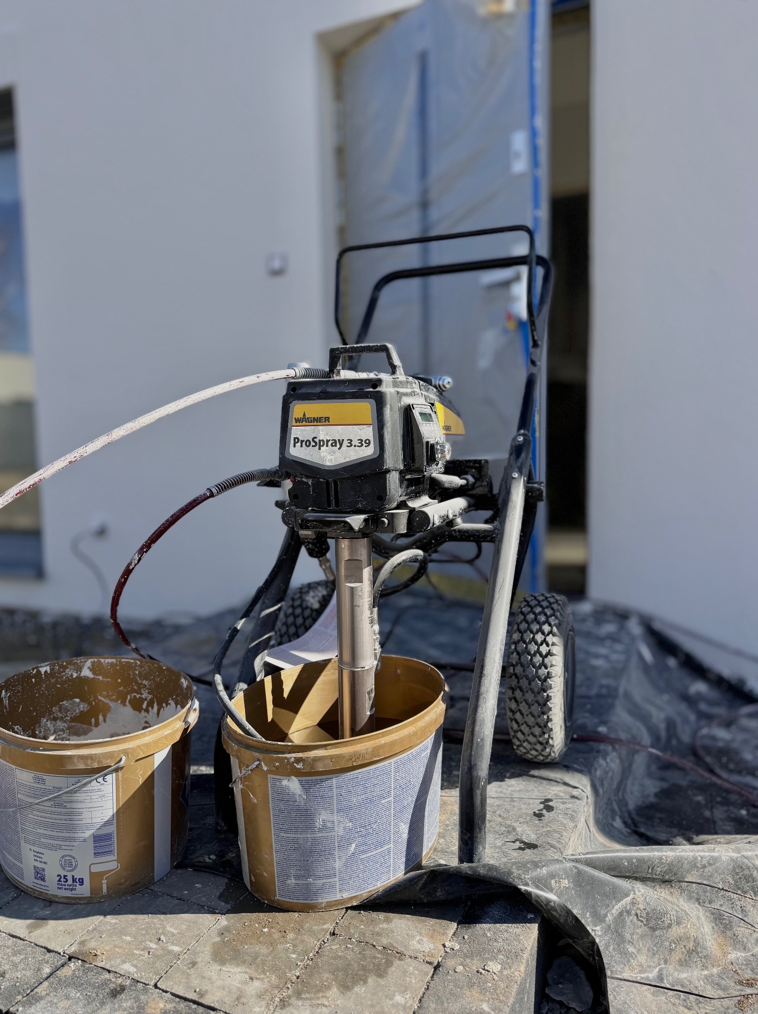 Profesjonalny agregat malarski Wagner ProSpray 3.39 z dwoma wiadrami farby na zewnątrz budynku w trakcie prac malarskich.