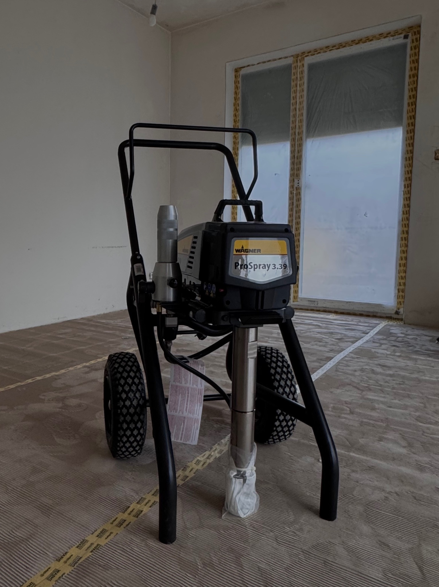 Profesjonalny agregat malarski Wagner ProSpray 3.39 na tle zabezpieczonego folią okna i przygotowanej do malowania podłogi w pomieszczeniu.