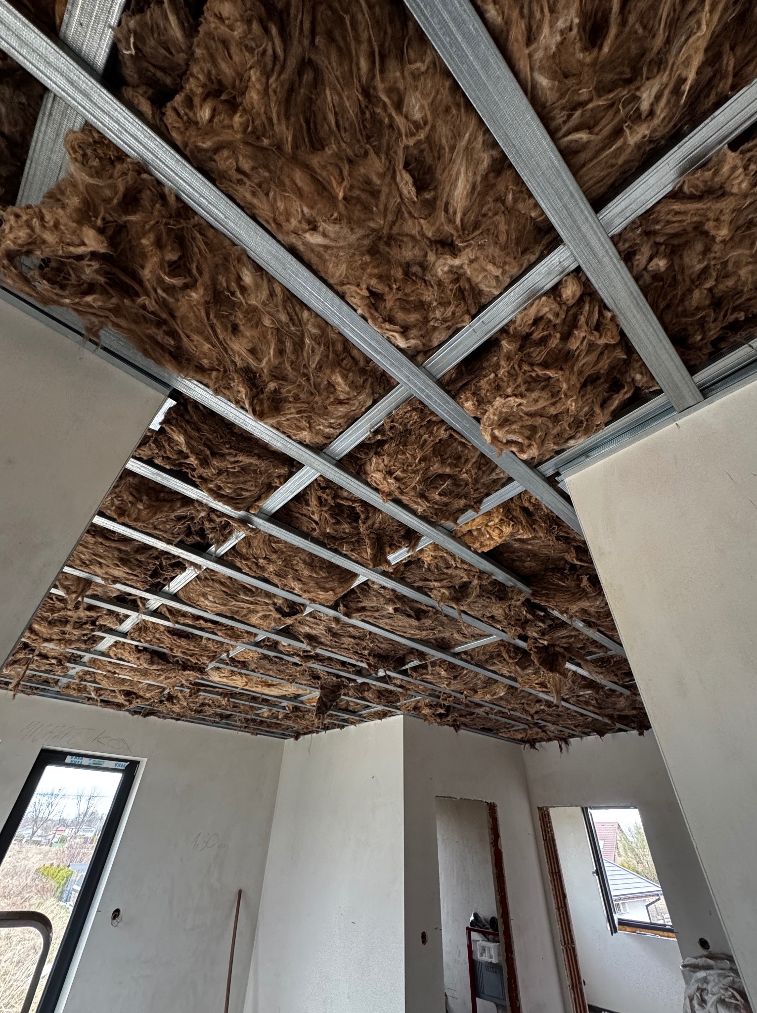 Sufit podwieszany z wełną mineralną w trakcie montażu, widoczne profile metalowe i izolacja, surowe ściany w tle, okna dachowe.