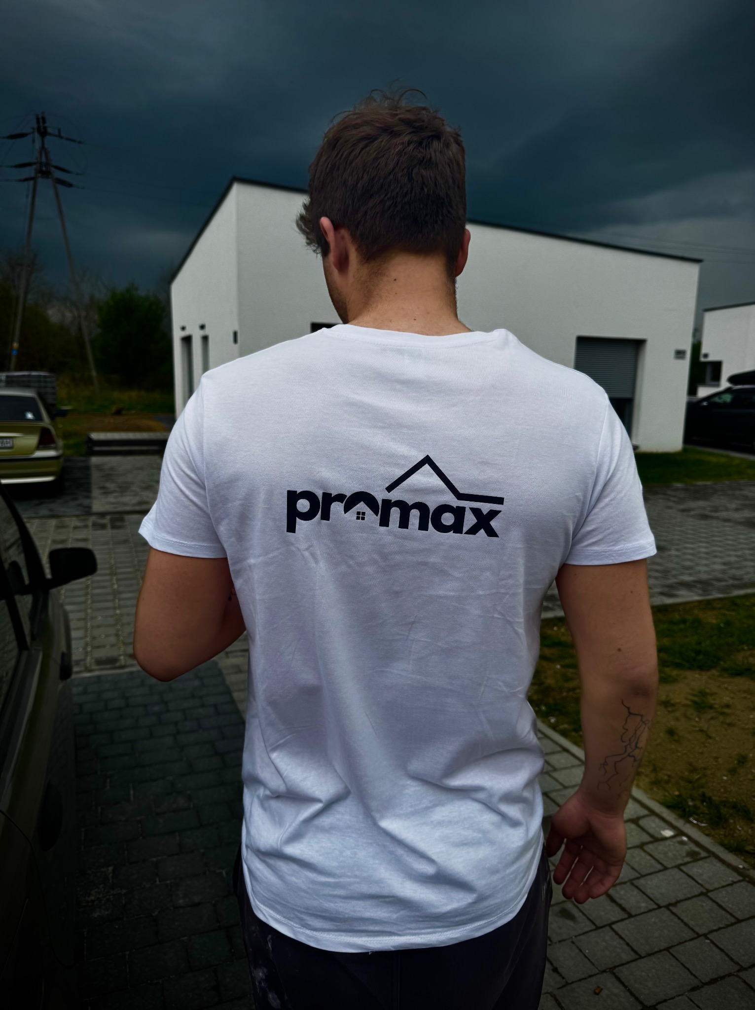 Mężczyzna w Żorach, tyłem, w białej koszulce z logo firmy budowlanej na tle nowoczesnego domu i burzowego nieba. Widoczny fragment samochodu i słup elektryczny.