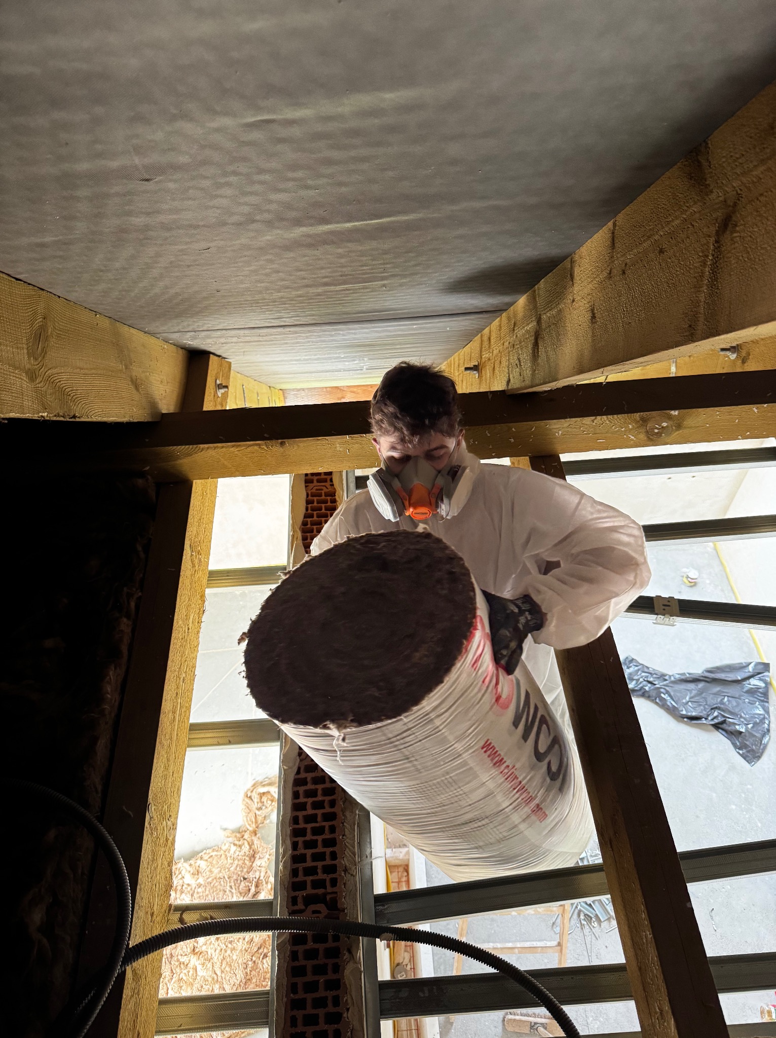 Pracownik w kombinezonie i masce trzyma rolkę wełny mineralnej na poddaszu podczas ocieplania, widoczne drewniane elementy konstrukcyjne i okno.