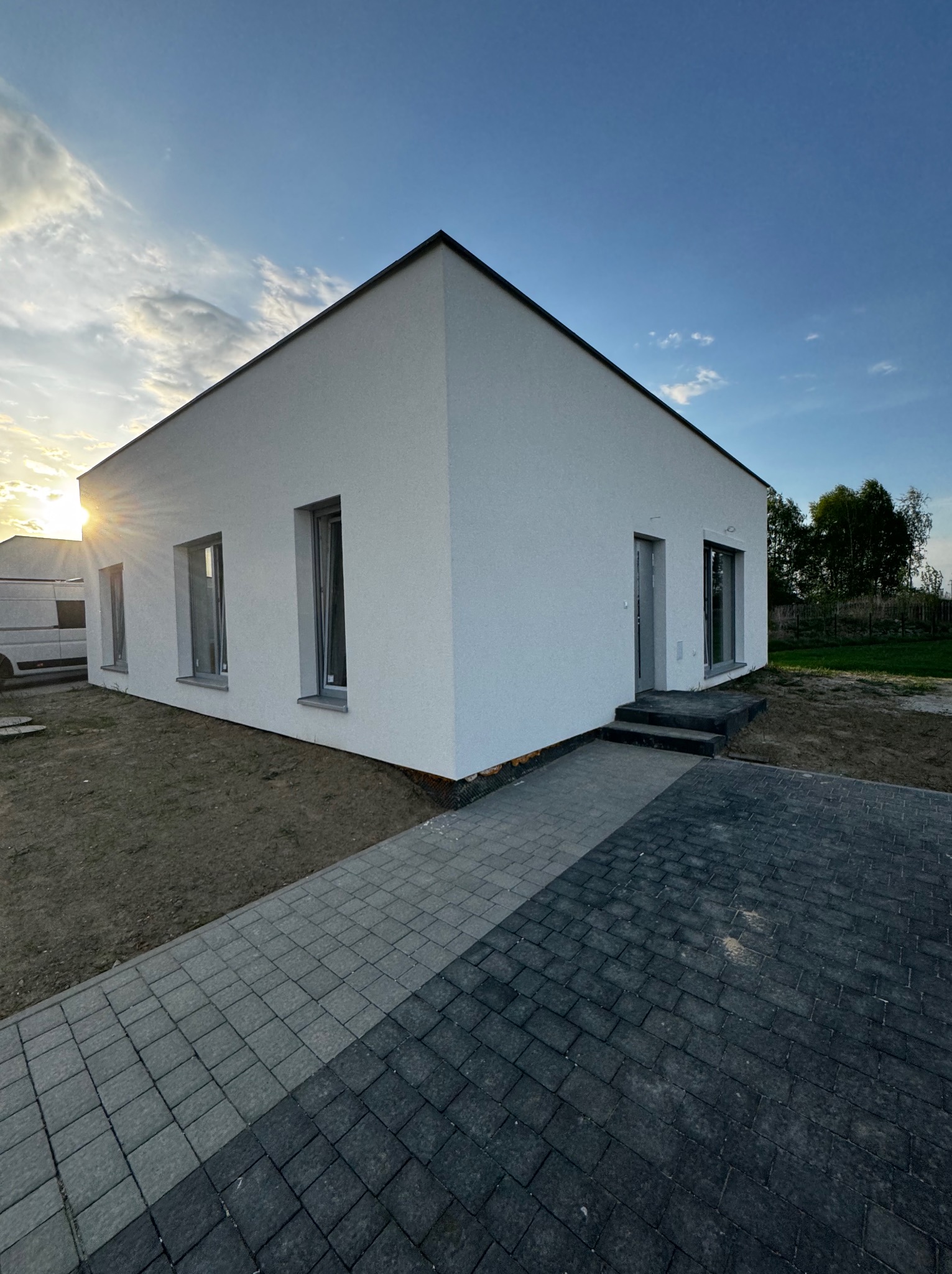 Nowoczesny, parterowy dom z białym tynkiem, szarą kostką brukową i trzema oknami. Widoczne drzwi wejściowe i fragment busa na tle zachodzącego słońca.