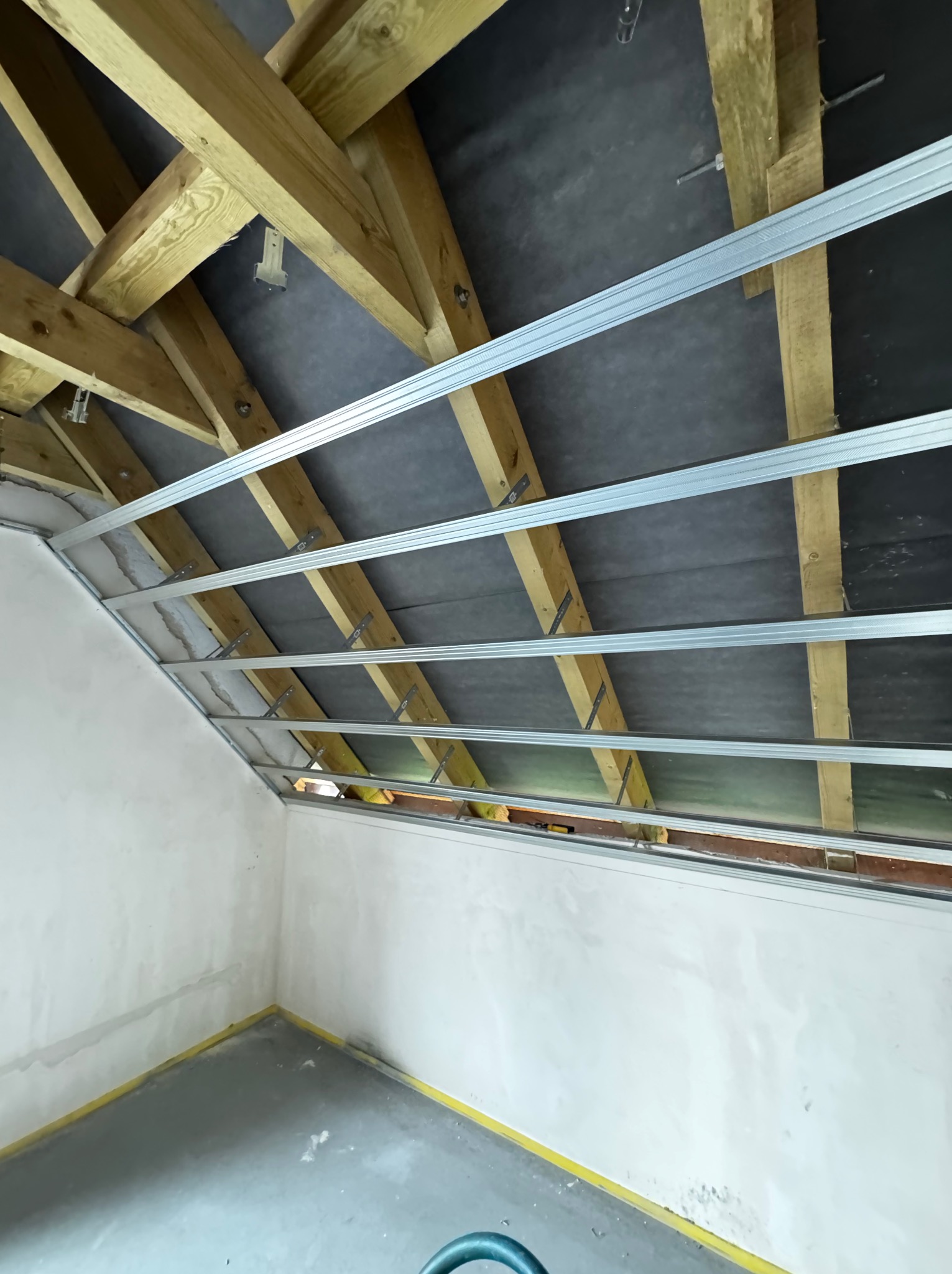 Szkielet pod płytę G-K na drewnianej konstrukcji dachu, widok z dołu. Metalowe profile zamocowane do drewnianych belek. Ściany i podłoga przygotowane do dalszych prac.