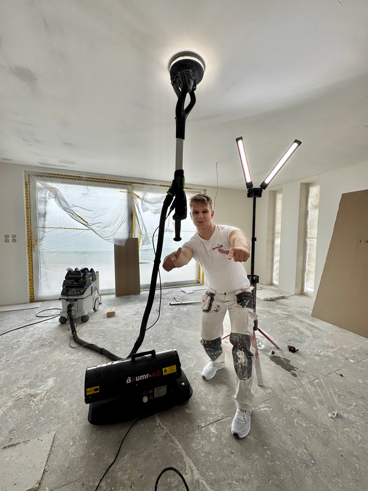 Mężczyzna z szlifierką do gładzi na wysięgniku, w trakcie pracy w jasnym pomieszczeniu. Widoczne lampy, okna zabezpieczone folią i odkurzacz przemysłowy.