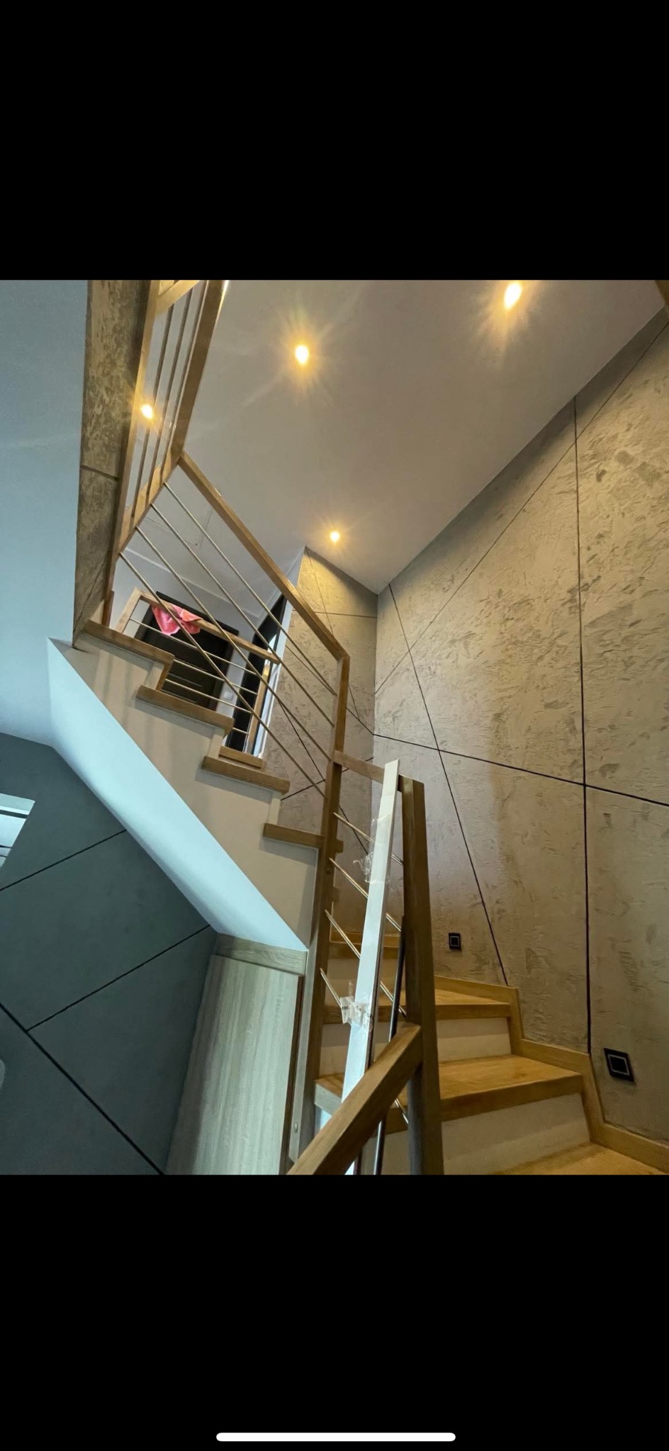 Nowoczesne schody drewniane z metalową balustradą w jasnym wnętrzu. Ściany wykończone w dekoracyjnym tynku z geometrycznym wzorem. Oświetlenie punktowe na suficie.