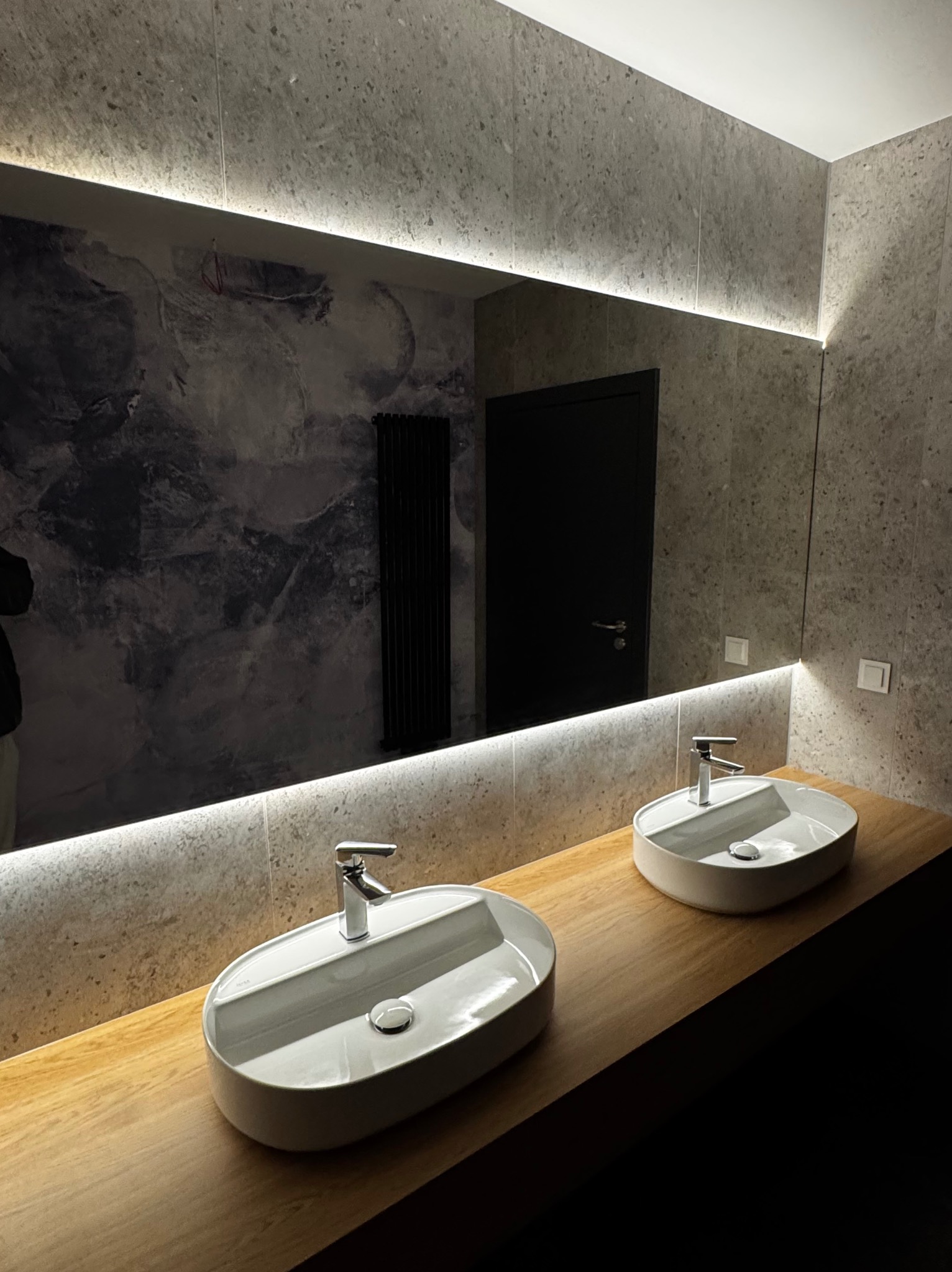 Nowoczesna łazienka z dwiema umywalkami na drewnianym blacie, podświetlane lustro na ścianie z efektem marmuru. Minimalistyczny design, ciemne drzwi w tle.