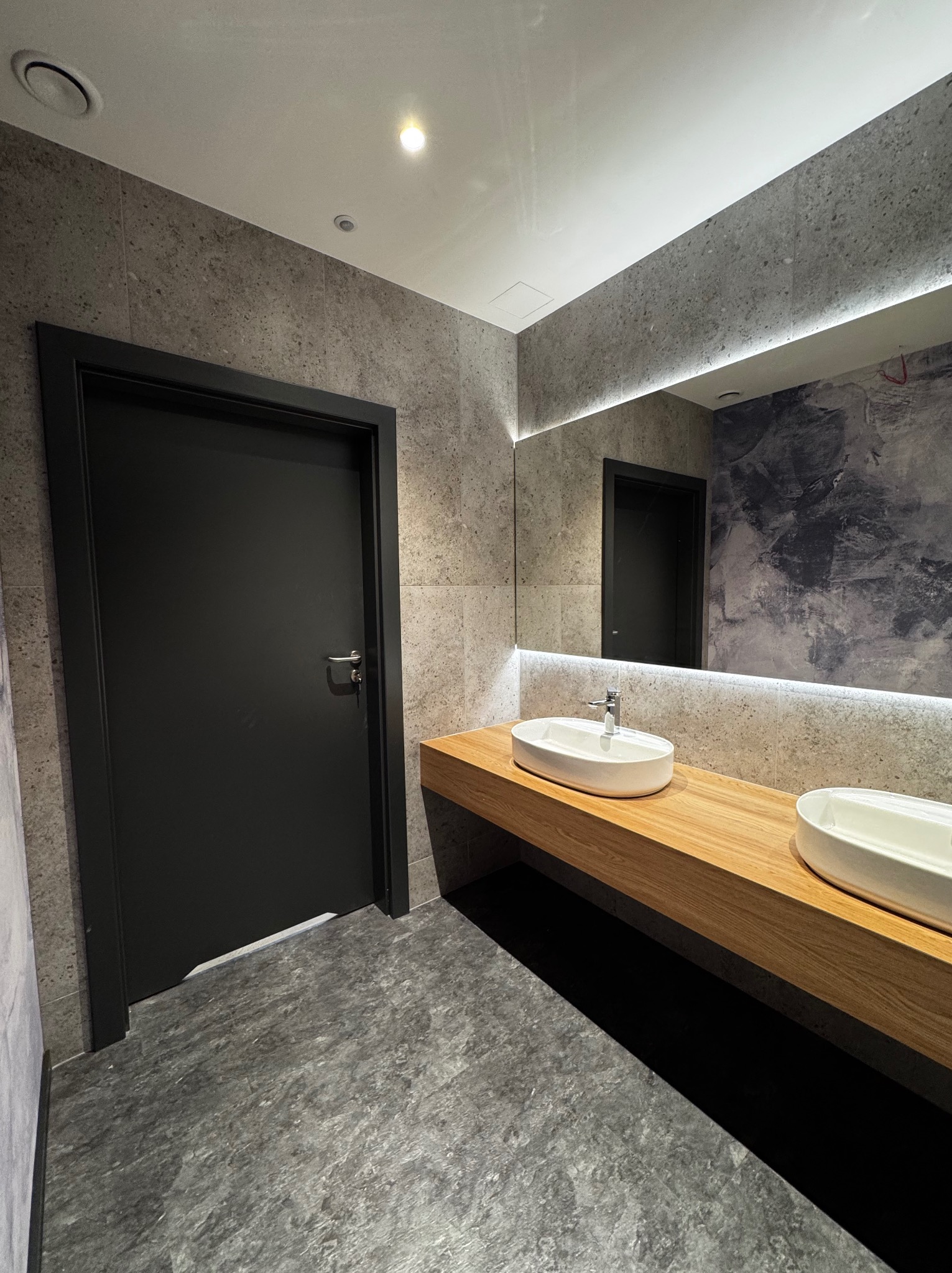 Nowoczesna łazienka z szarą okładziną, drewnianym blatem pod umywalki i czarnymi drzwiami. Oświetlenie LED podkreśla lustro i sufit.