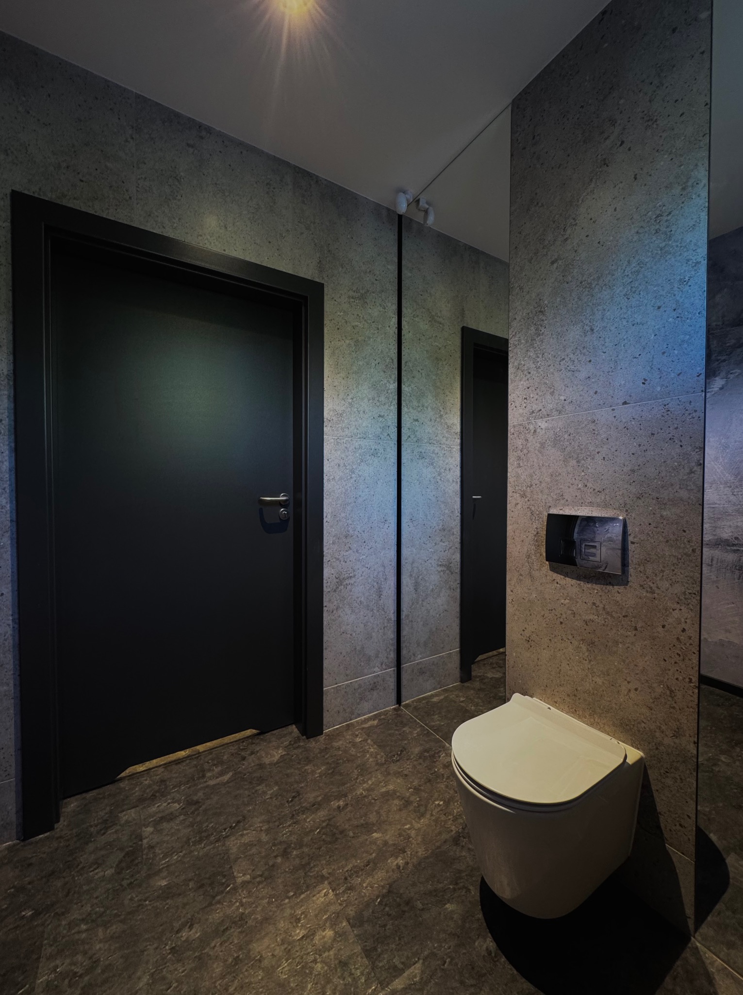 Nowoczesna łazienka z czarnymi drzwiami, szarą płytką imitującą kamień i podwieszaną toaletą. Lustro optycznie powiększa przestrzeń. Minimalistyczny design.