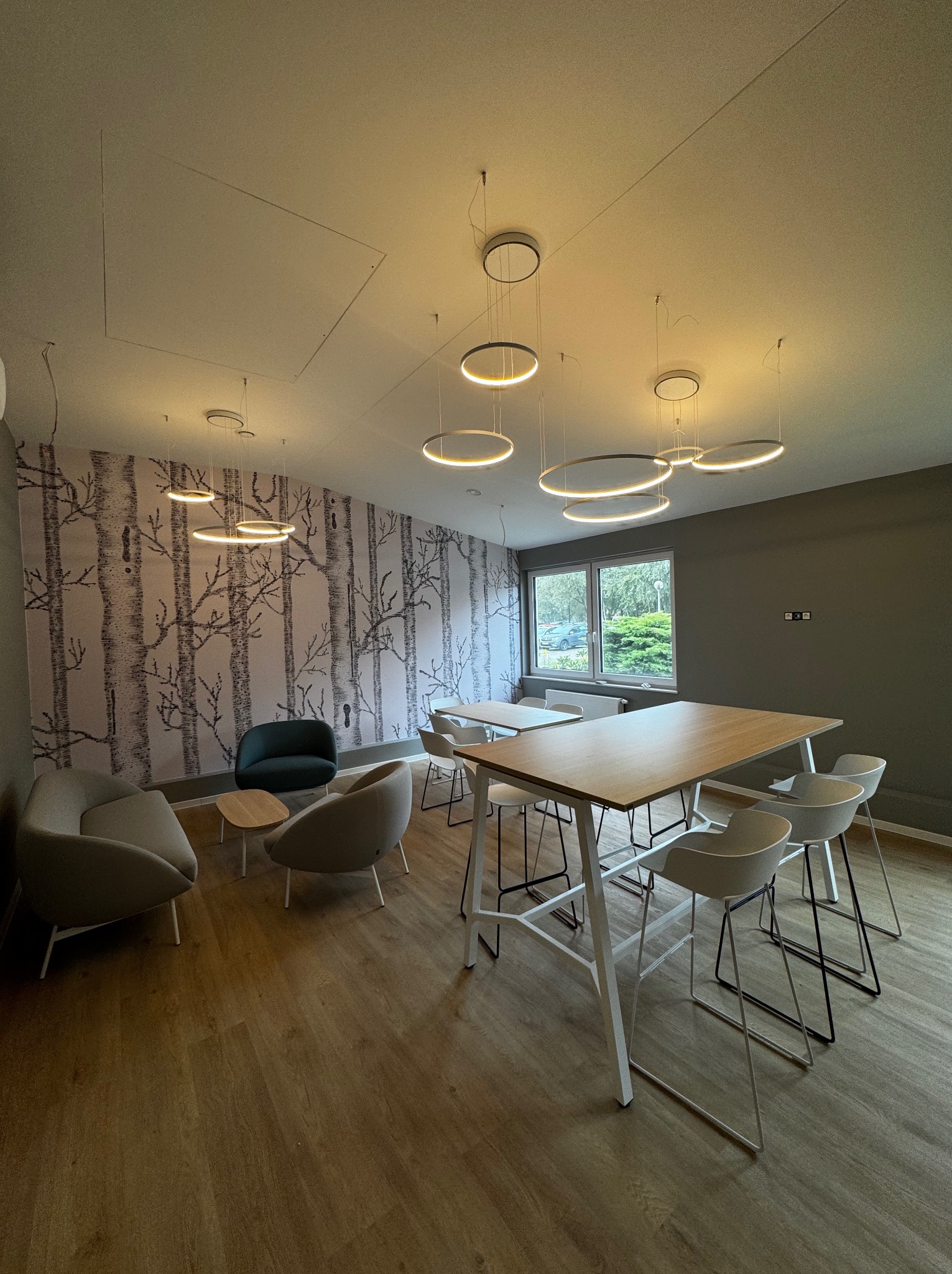 Nowoczesne wnętrze biurowe z tapetą w motywie drzew, okrągłymi lampami i drewnianą podłogą. Widoczne stoły z krzesłami i strefa relaksu z fotelami.