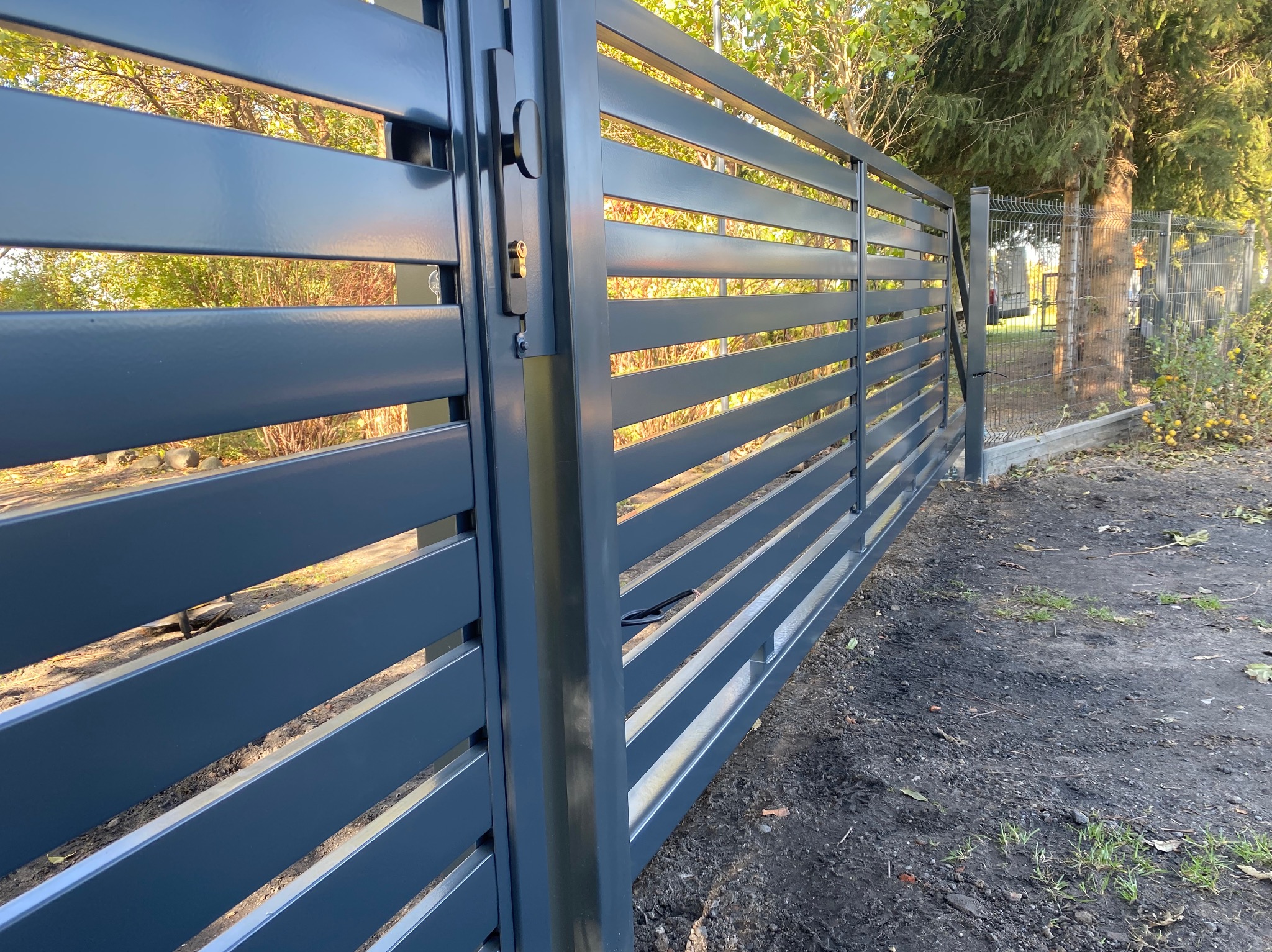 Nowoczesna przesuwna brama w kolorze grafitowym z poziomymi panelami, widoczny zamek i otoczenie z drzewami i siatką ogrodzeniową.