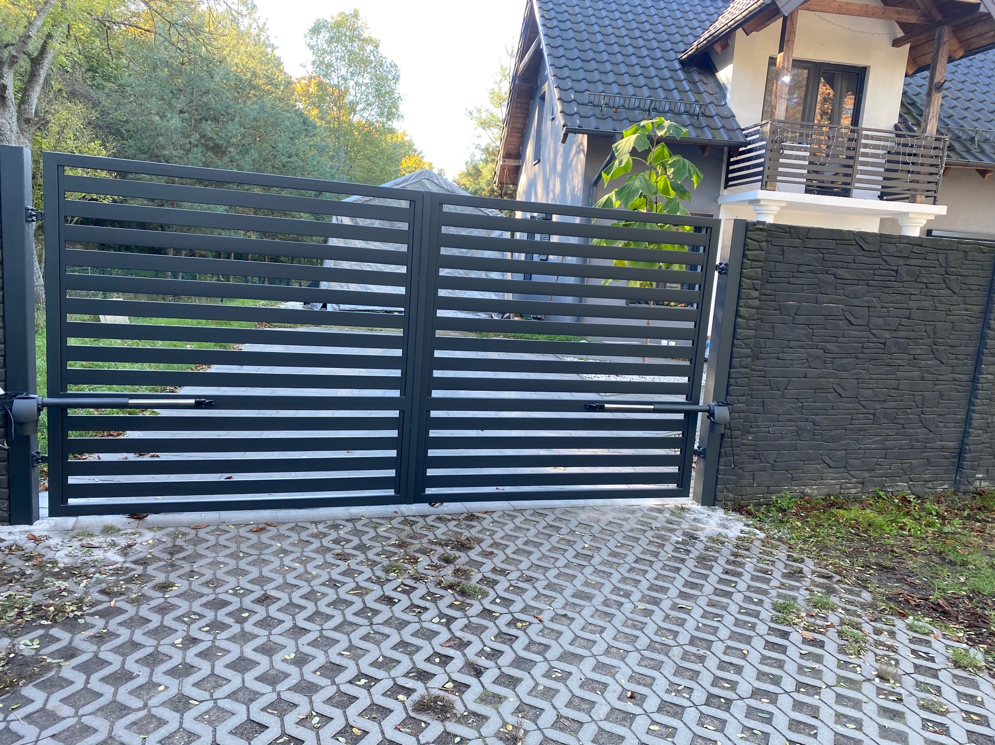 Nowoczesna, pozioma brama dwuskrzydłowa w kolorze antracytowym, z automatycznym napędem, na tle domu z balkonem i kostki brukowej.