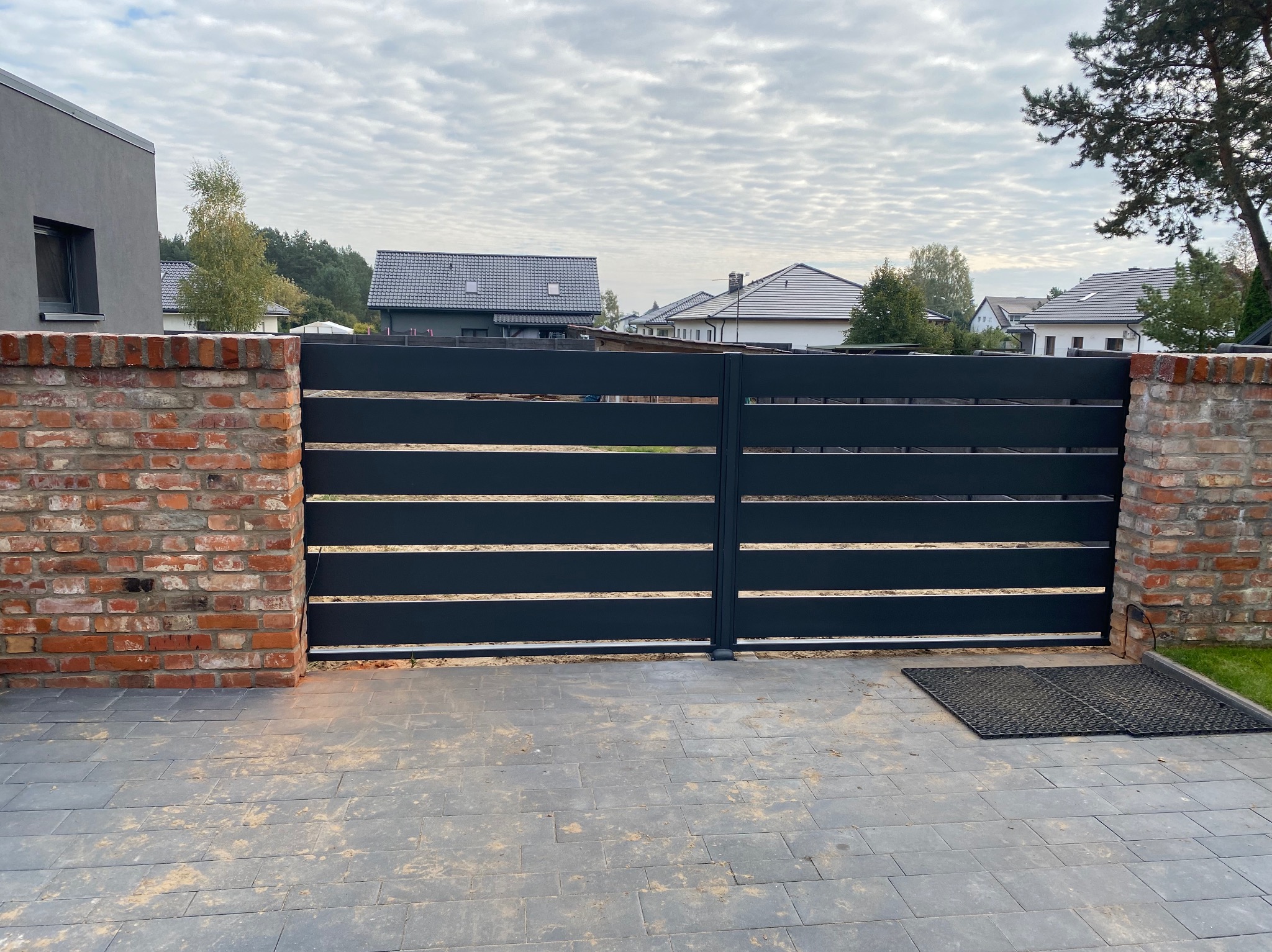 Nowoczesna, pozioma brama wjazdowa z czarnych paneli, osadzona między ceglanymi słupkami, na tle brukowanej nawierzchni i zabudowy jednorodzinnej.