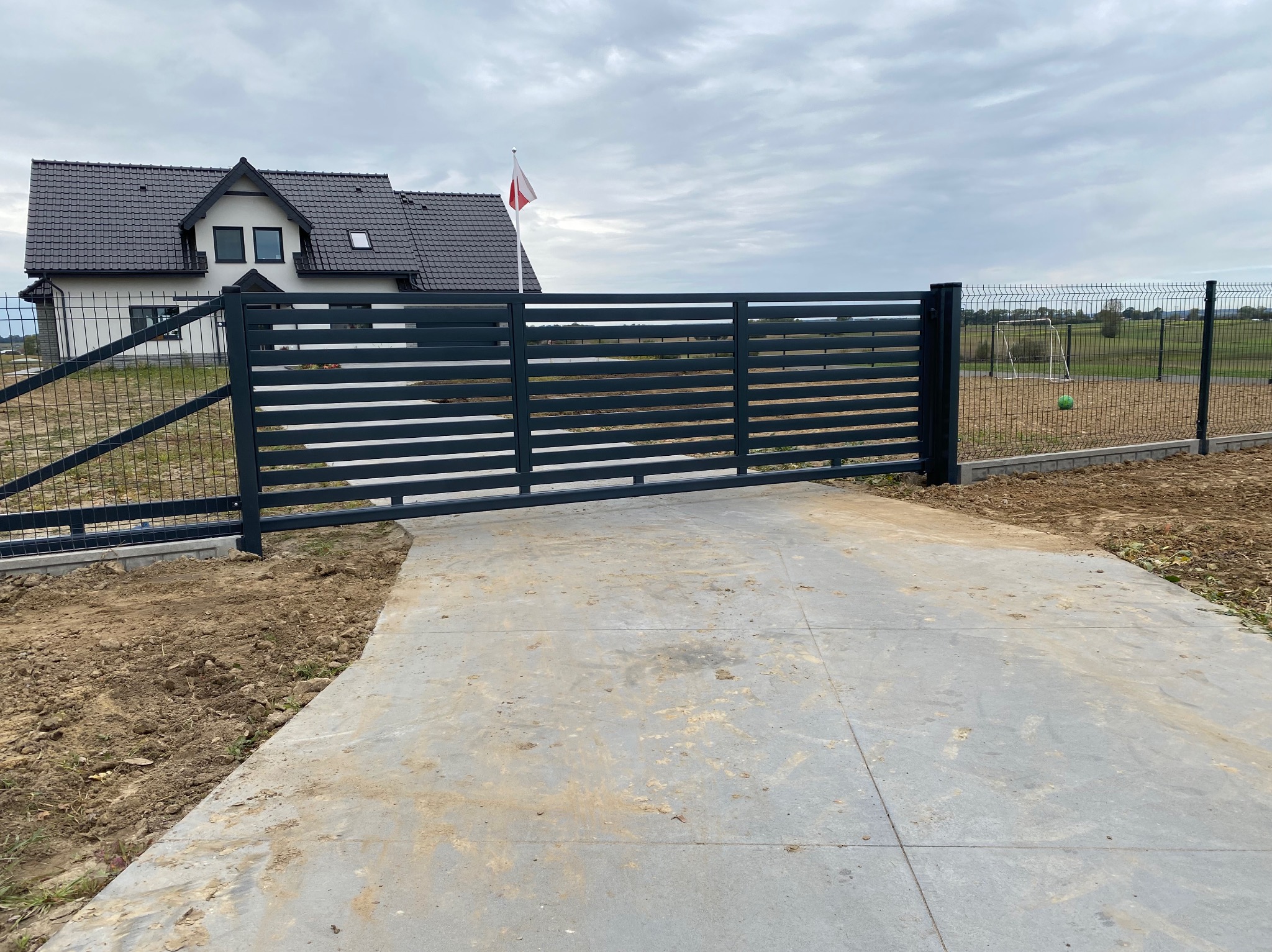 Nowoczesna, przesuwna brama w kolorze antracytu z poziomymi panelami, widoczna na tle domu z polską flagą i siatki ogrodzeniowej.