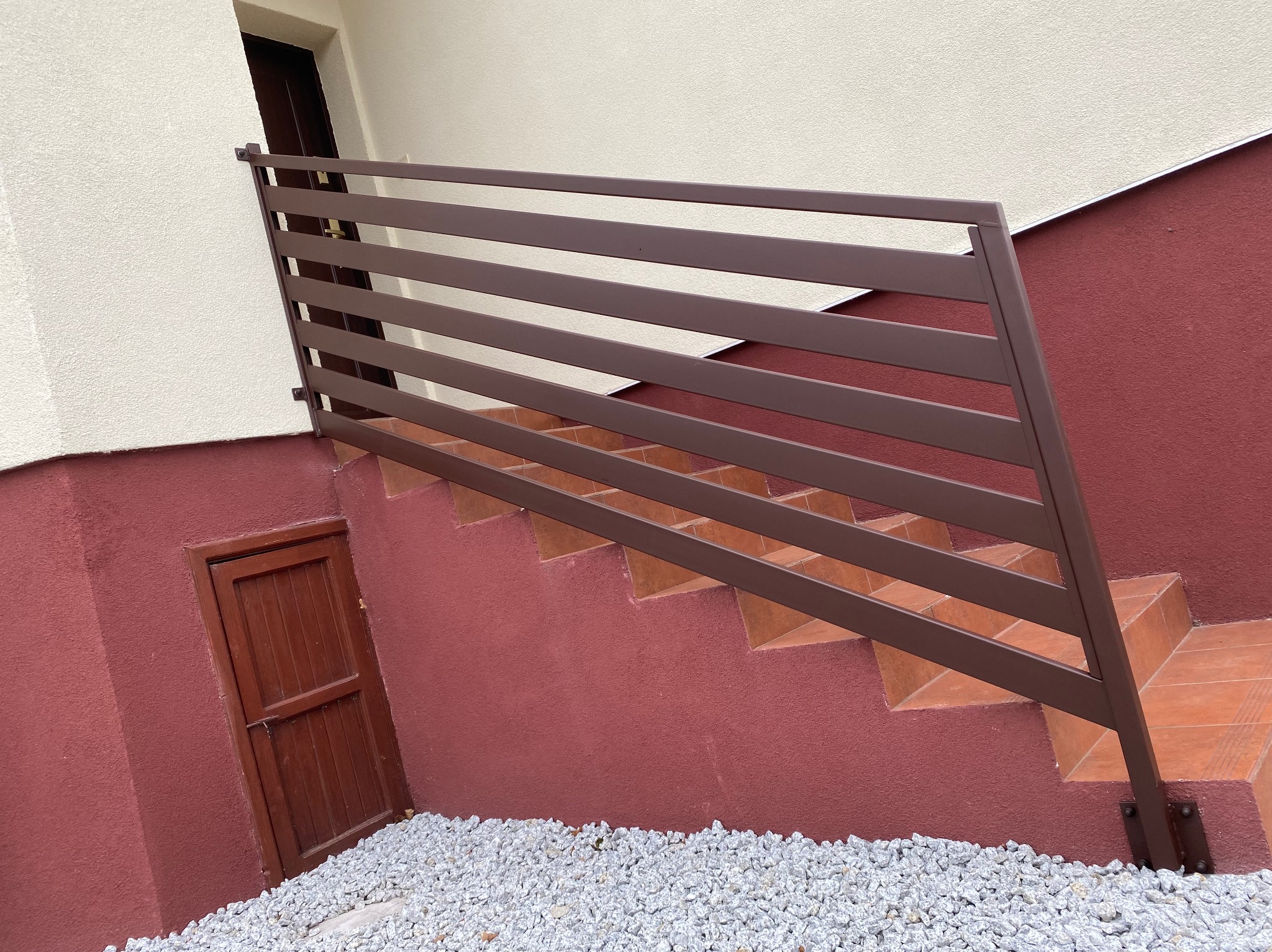 Brązowa, pozioma balustrada z profili na schodach zewnętrznych, prowadzących do drzwi piwnicznych, z widocznym podsypaniem z białego kamienia.