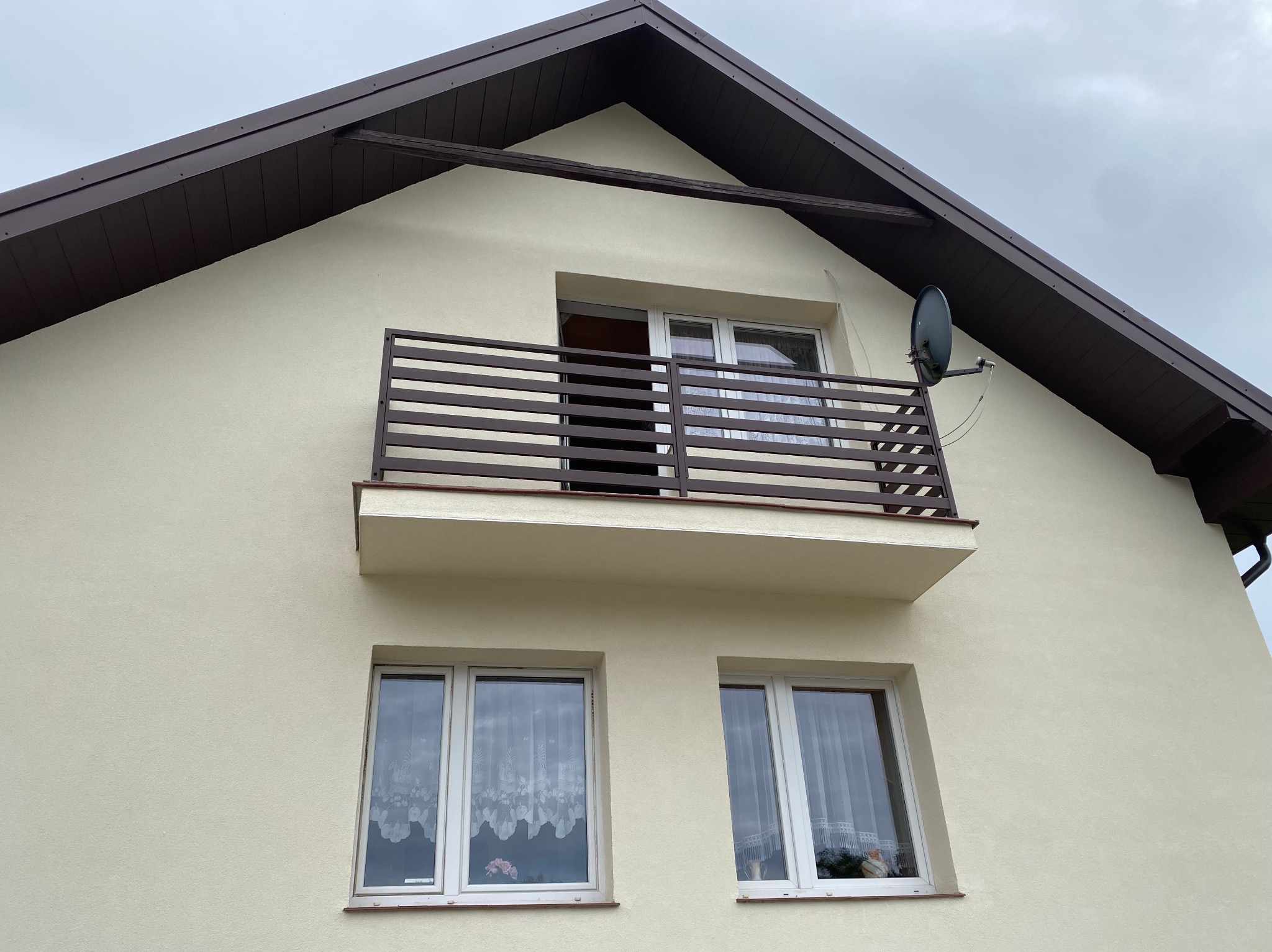 Jasna elewacja domu z balkonem z brązową, poziomą balustradą, z widoczną anteną satelitarną po prawej stronie i oknami z białymi ramami oraz firankami na parterze.