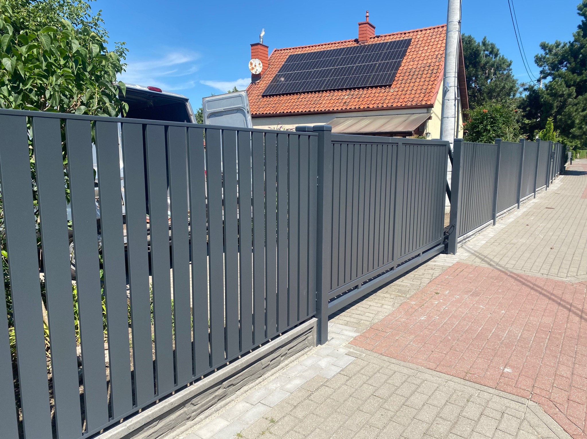 Nowoczesna, szara brama przesuwna z pionowymi panelami, widoczna na tle domu z panelami słonecznymi i zaparkowanym samochodem dostawczym.