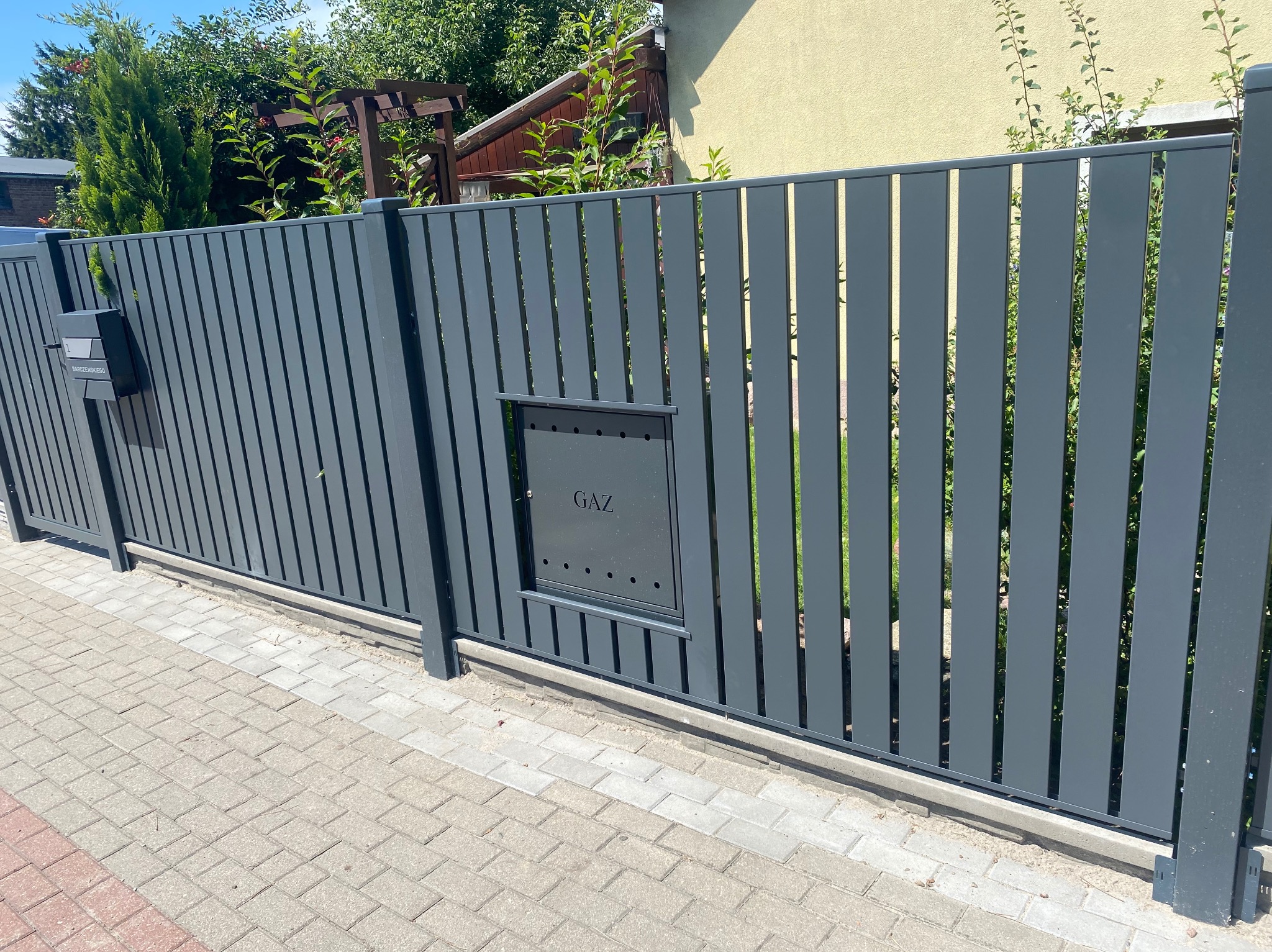 Nowoczesna, grafitowa brama posesyjna z pionowymi panelami i skrzynką gazową wbudowaną w konstrukcję, widoczna z poziomu chodnika z kostki brukowej.