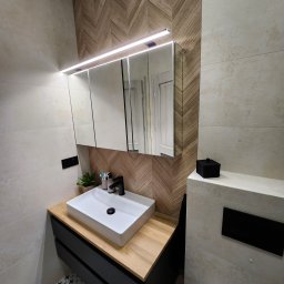 MAT-DACH BUD - Nowoczesna łazienka z umywalką nablatową, czarną armaturą i drewnopodobną ścianą w jodełkę, oświetlona ledowym światłem nad lustrem.