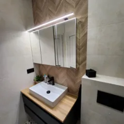 Nowoczesna łazienka z umywalką nablatową, czarną armaturą i drewnopodobną ścianą w jodełkę, oświetlona ledowym światłem nad lustrem.