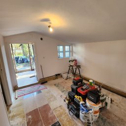 MAT-DACH BUD - Wnętrze pomieszczenia w trakcie remontu, widoczne narzędzia, wiadra z zaprawą, drabina, podłoga z płytek w różnych kolorach, ściany przygotowane do malowania, otwarte drzwi na zewnątrz.