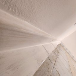 MAT-DACH BUD - Niedokładnie pomalowany narożnik ściany i sufitu z widocznymi smugami farby i zabrudzeniami na podłodze z płytek.