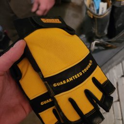 MAT-DACH BUD - Żółto-czarne rękawice robocze marki DeWalt, używane przez fachowca z Kołobrzegu, z widocznym napisem 'GUARANTEED TOUGH', trzymane w dłoni na tle ściany i narzędzi.