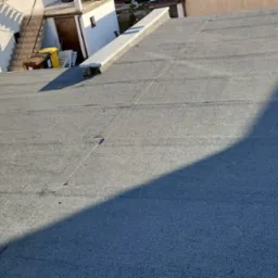 Widok z góry na szary, płaski dach pokryty papą termozgrzewalną, z widocznym kominem wentylacyjnym i fragmentem budynku w tle.