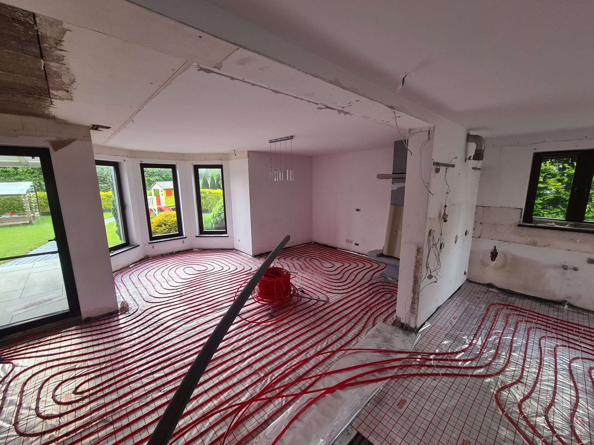 Rozłożone czerwone rury ogrzewania podłogowego na folii izolacyjnej w surowym wnętrzu domu przed wylewką, widoczne okna z widokiem na ogród i niedokończone ściany.