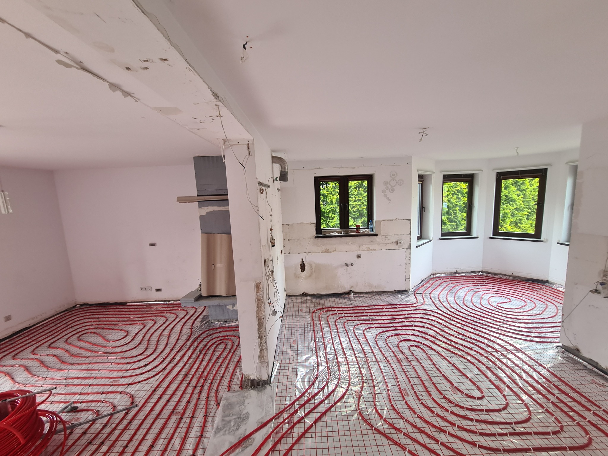 Instalacja ogrzewania podłogowego z czerwonych rurek na folii w pomieszczeniu w trakcie remontu, widoczne ściany w stanie surowym i okna z zielenią na zewnątrz.