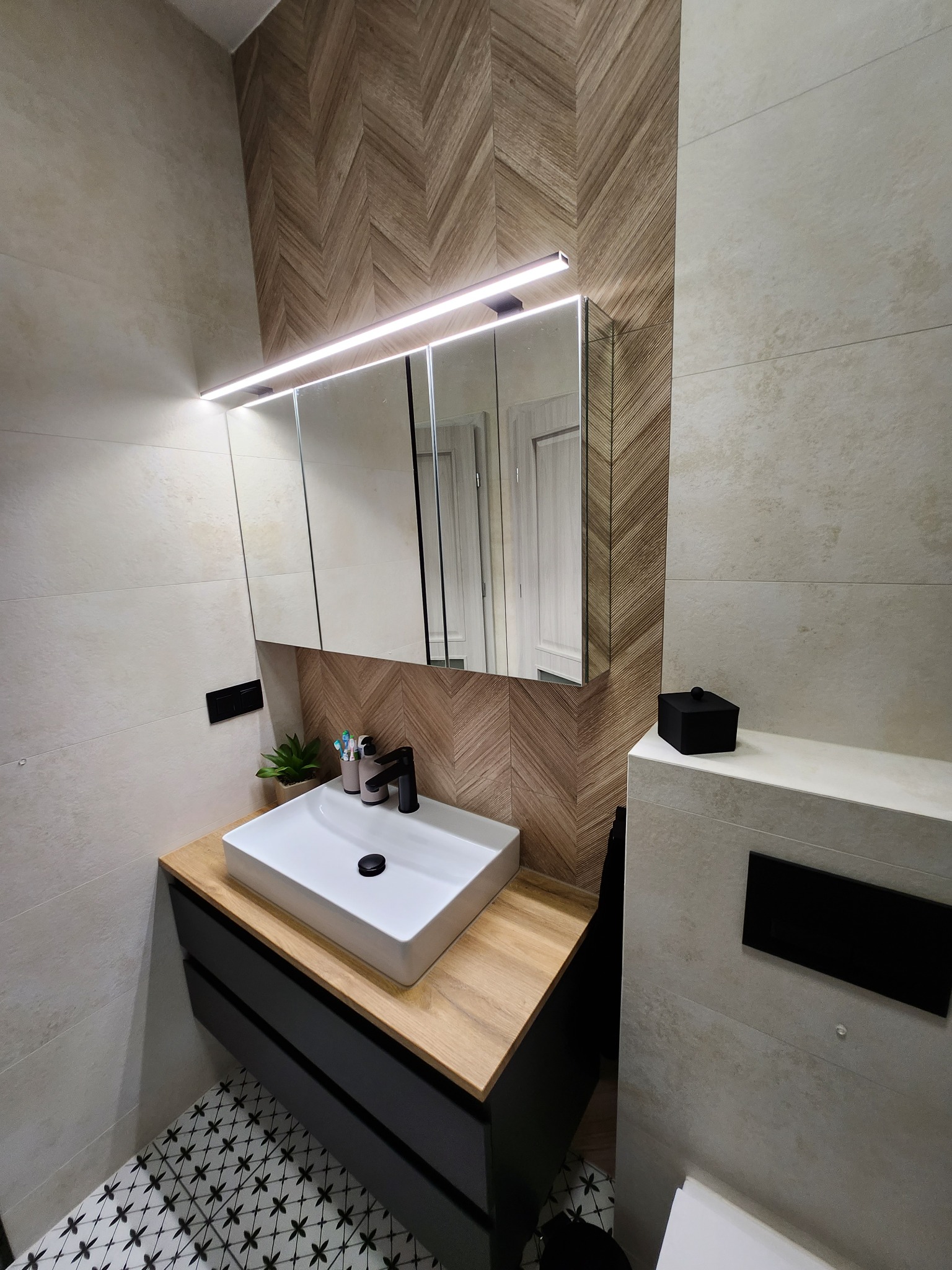 Nowoczesna łazienka z umywalką nablatową, czarną armaturą i drewnopodobną ścianą w jodełkę, oświetlona ledowym światłem nad lustrem.