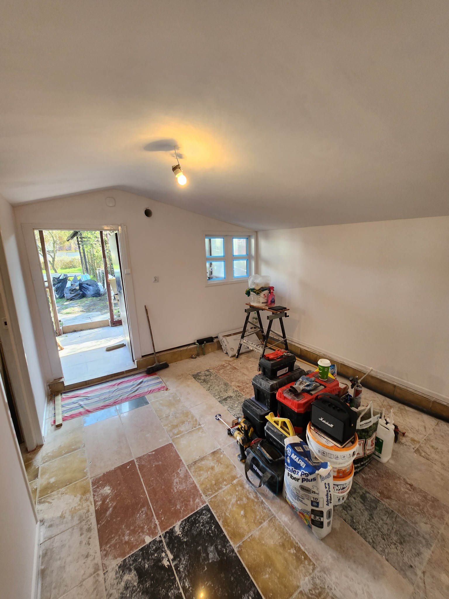 Wnętrze pomieszczenia w trakcie remontu, widoczne narzędzia, wiadra z zaprawą, drabina, podłoga z płytek w różnych kolorach, ściany przygotowane do malowania, otwarte drzwi na zewnątrz.