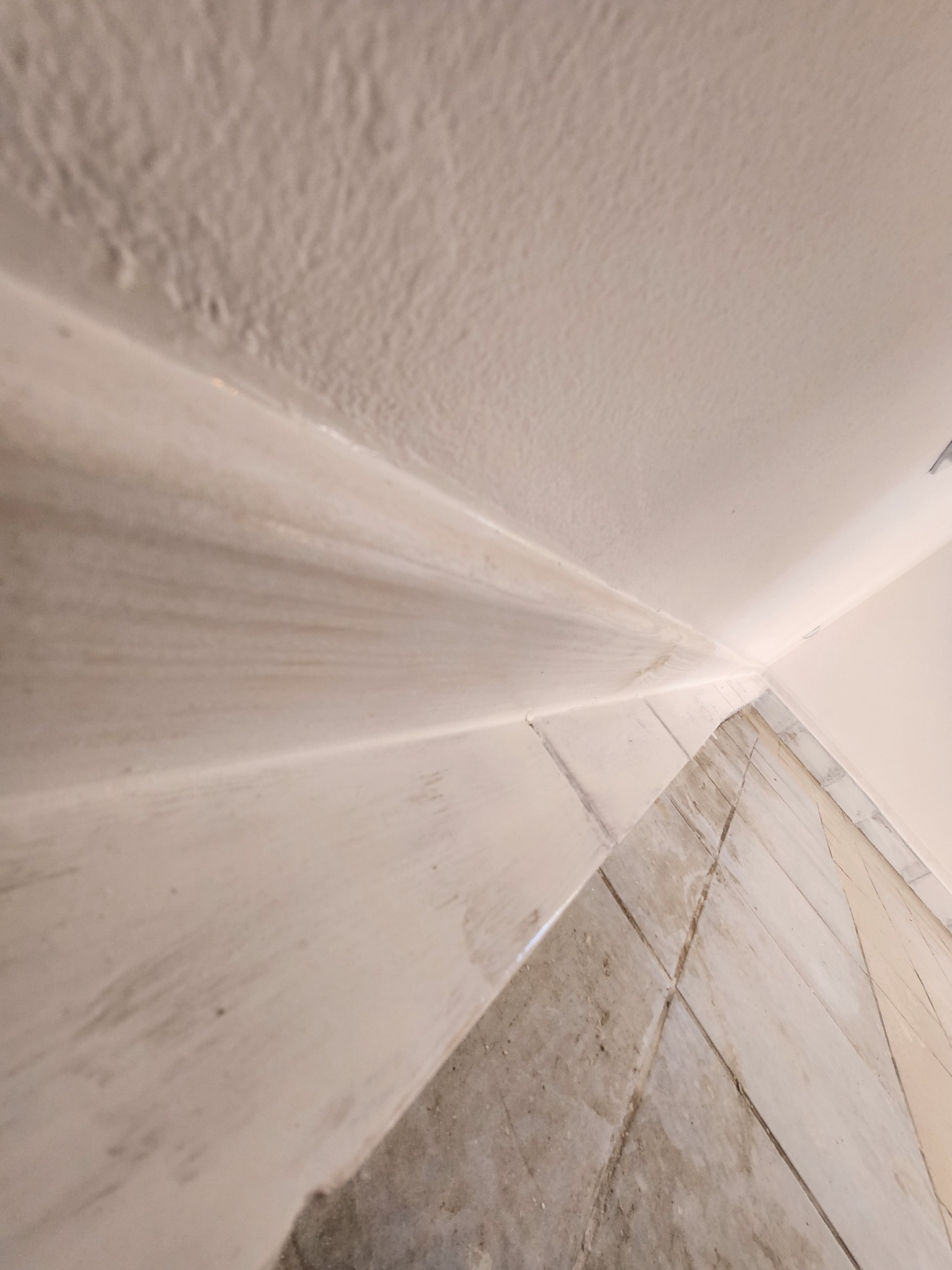 Niedokładnie pomalowany narożnik ściany i sufitu z widocznymi smugami farby i zabrudzeniami na podłodze z płytek.