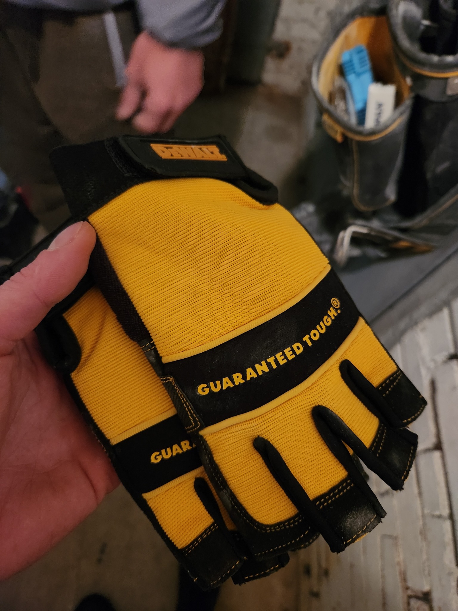 Żółto-czarne rękawice robocze marki DeWalt, używane przez fachowca z Kołobrzegu, z widocznym napisem 'GUARANTEED TOUGH', trzymane w dłoni na tle ściany i narzędzi.