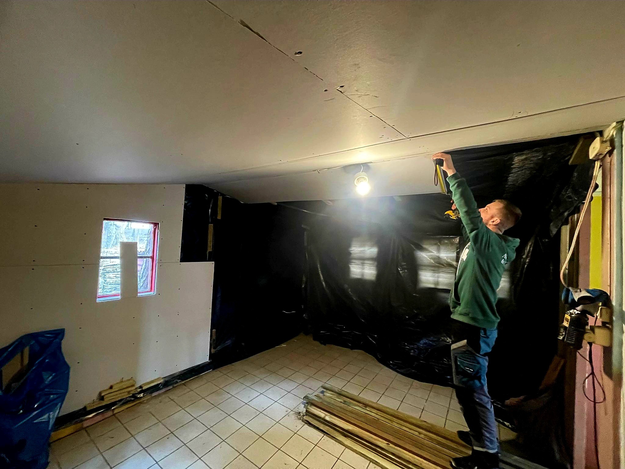 Mężczyzna w zielonej bluzie mierzy sufit z płyt kartonowo-gipsowych w pomieszczeniu zabezpieczonym folią, widoczne deski i narzędzia.