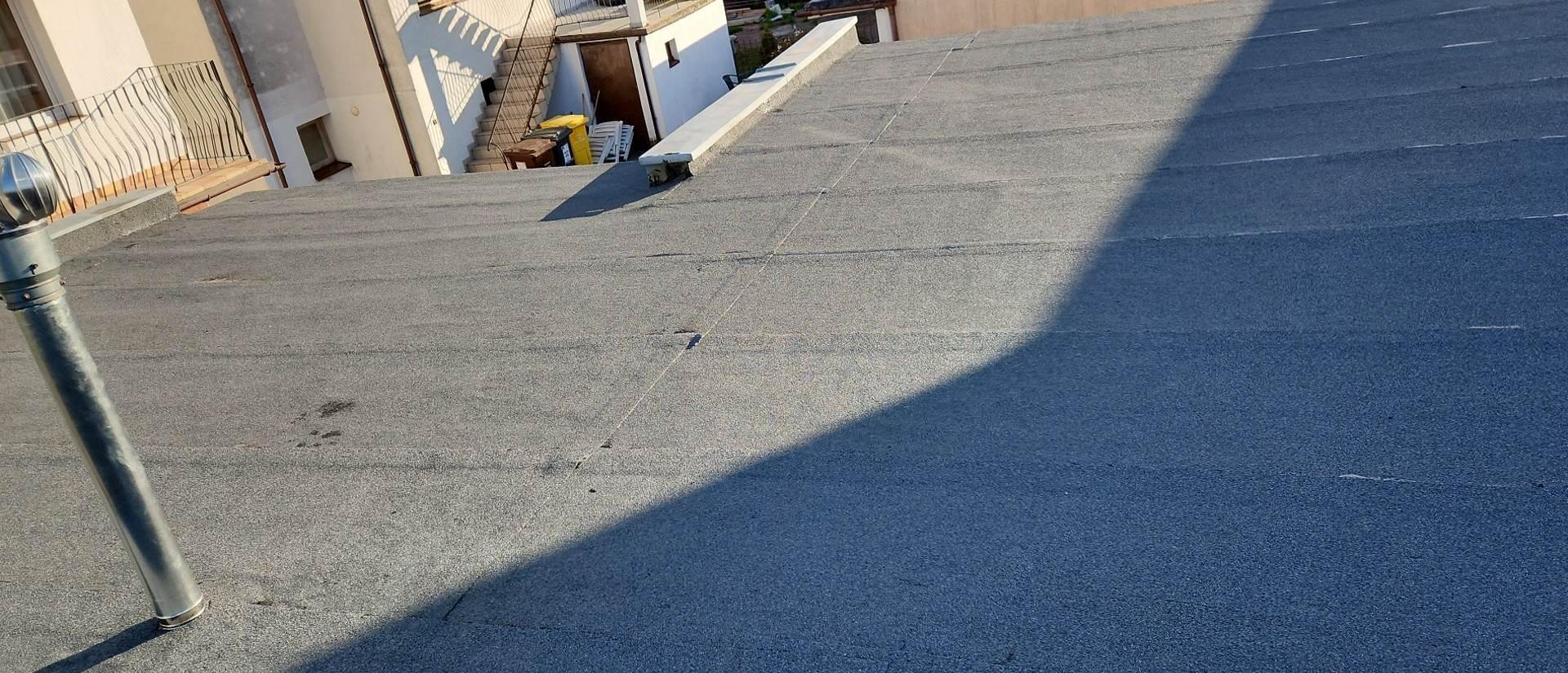 Widok z góry na szary, płaski dach pokryty papą termozgrzewalną, z widocznym kominem wentylacyjnym i fragmentem budynku w tle.