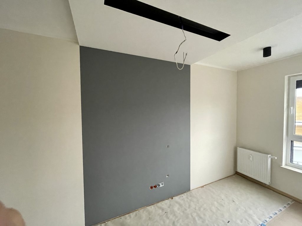 Pokój po malowaniu: jedna ściana szara, druga kremowa, zabezpieczona podłoga, instalacja elektryczna i oświetleniowa w trakcie montażu.