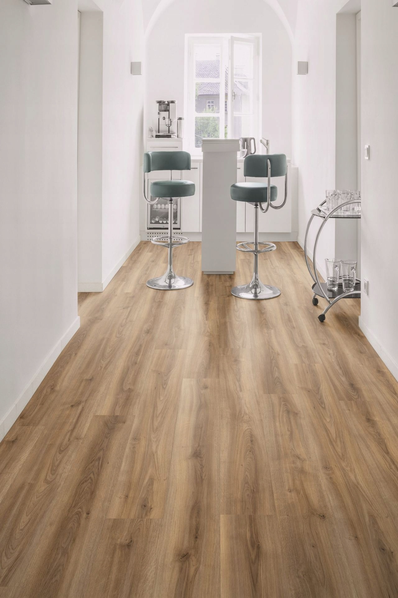 Jasnobrązowe panele podłogowe imitujące drewno, ułożone wzdłużnie w nowoczesnej, białej kuchni z barkiem, wysokimi krzesłami i przeszklonym wózkiem na napoje.