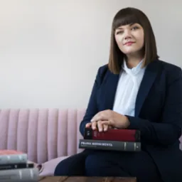 Portret kobiety prawnika w granatowym garniturze, siedzącej na różowej sofie z książkami, w tym 'Prawa Konsumenta', na drewnianym stoliku.