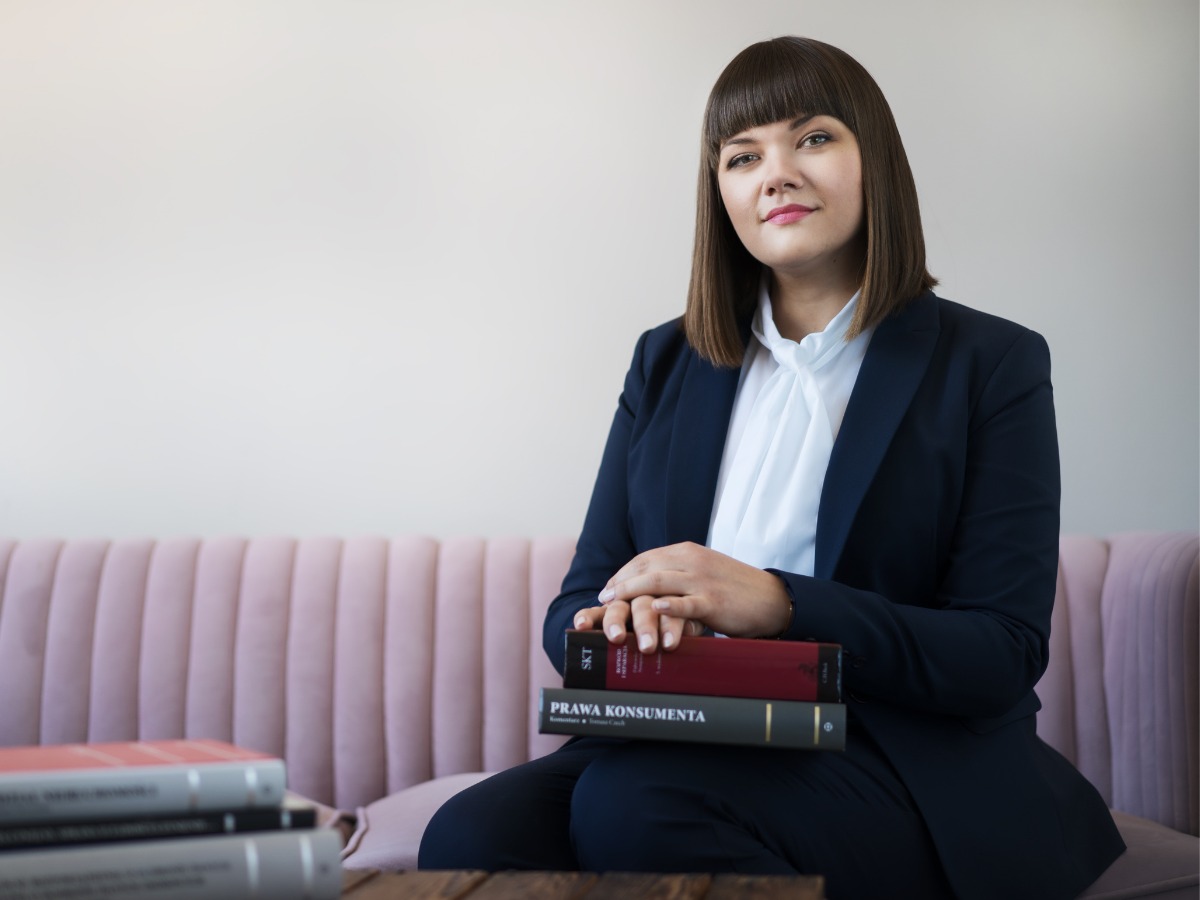 Portret kobiety prawnika w granatowym garniturze, siedzącej na różowej sofie z książkami, w tym 'Prawa Konsumenta', na drewnianym stoliku.
