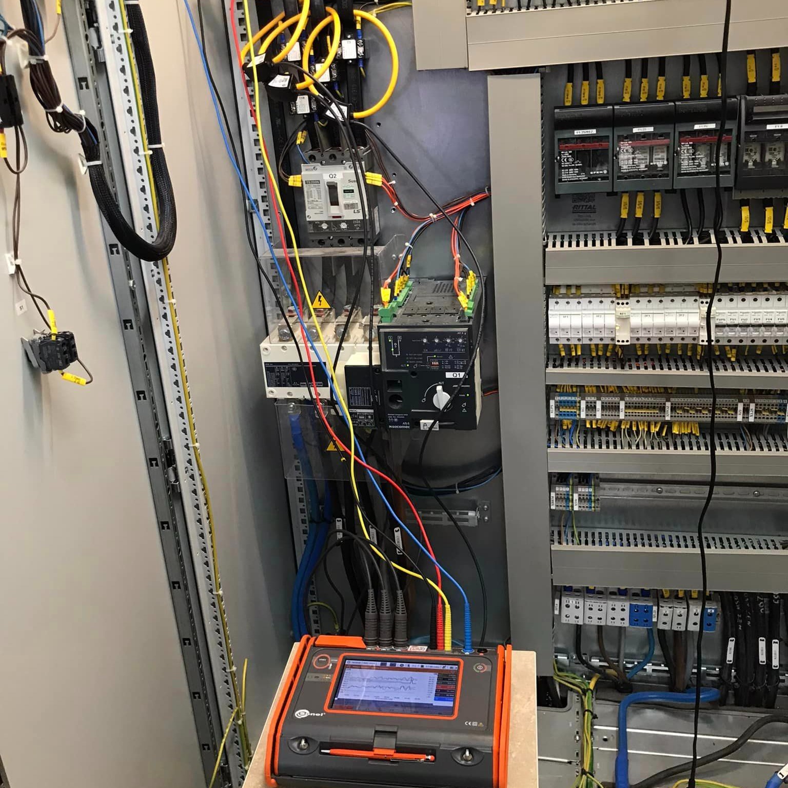 Panel elektryczny z licznymi przewodami i zabezpieczeniami, analizator jakości z widocznym wykresem pomiarowym, kable pomiarowe podłączone do urządzeń.