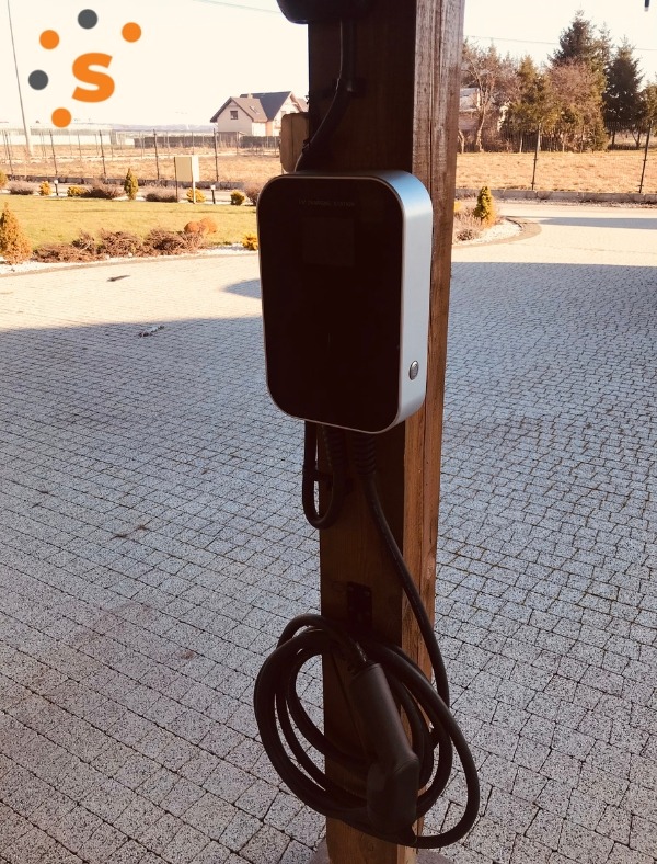 Szara stacja ładowania pojazdów elektrycznych zamontowana na drewnianym słupie, z kablem zwiniętym u dołu, na tle brukowanej nawierzchni i zieleni ogrodu.