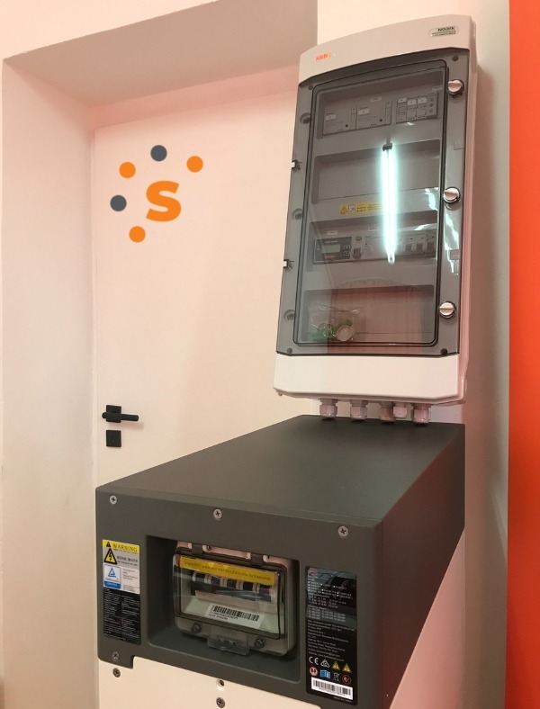 Rozdzielnica elektryczna z osprzętem zabezpieczającym, zamontowana na ścianie wewnątrz budynku, widoczny panel z bezpiecznikami i okablowaniem, obok logo firmy na ścianie.