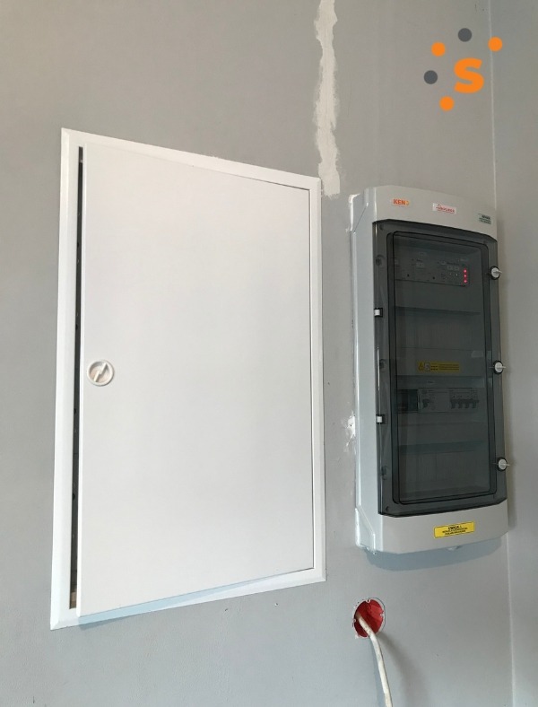 Szara ściana z zamontowaną białą skrzynką rozdzielczą i szarą szafką elektryczną z przezroczystymi drzwiczkami, widoczny wystający przewód elektryczny ze ściany.