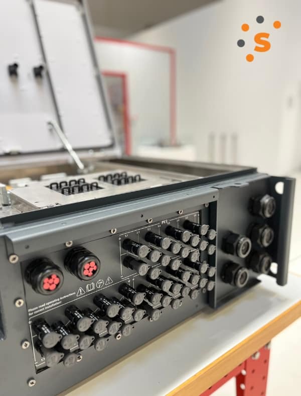 Otwarta skrzynka elektryczna z licznymi złączami i przewodami, widoczne oznaczenia i instrukcje bezpieczeństwa, w tle jasne pomieszczenie.